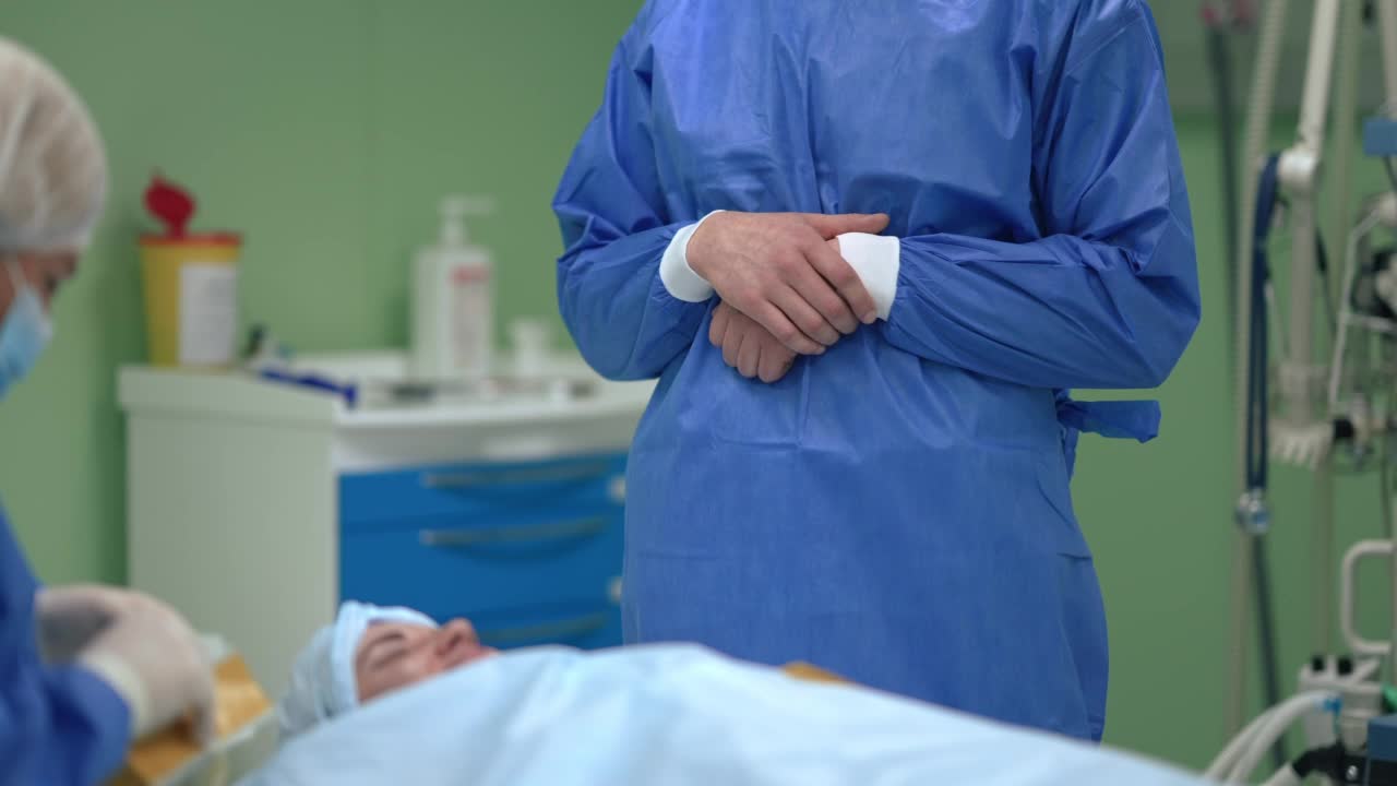 一名身穿外科制服的白人男子站在手术室里，以慢镜头为病人做准备。前方外科医生在医院室内等待护士协助。视频素材