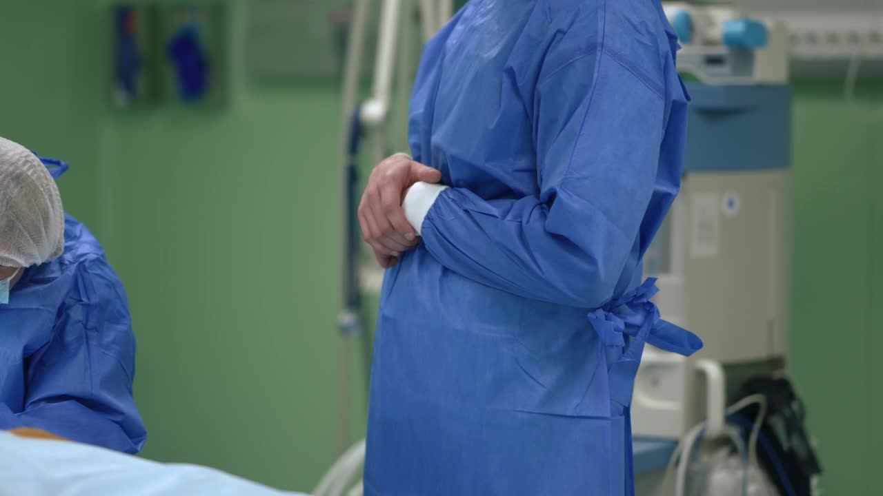 侧面图，不知名的外科医生穿着手术服，交叉双手站在手术室，护士为病人做准备。等待手术的白人男子从室内开始。缓慢的运动。视频素材