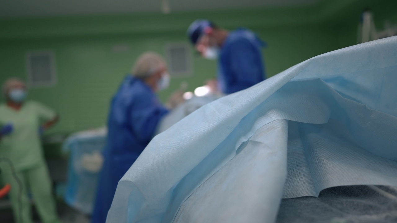 手术床上蓝色床单的特写，背景是模糊的团队手术病人。无法辨认的白人外科医生和助手在医院室内做手术。缓慢的运动。视频素材