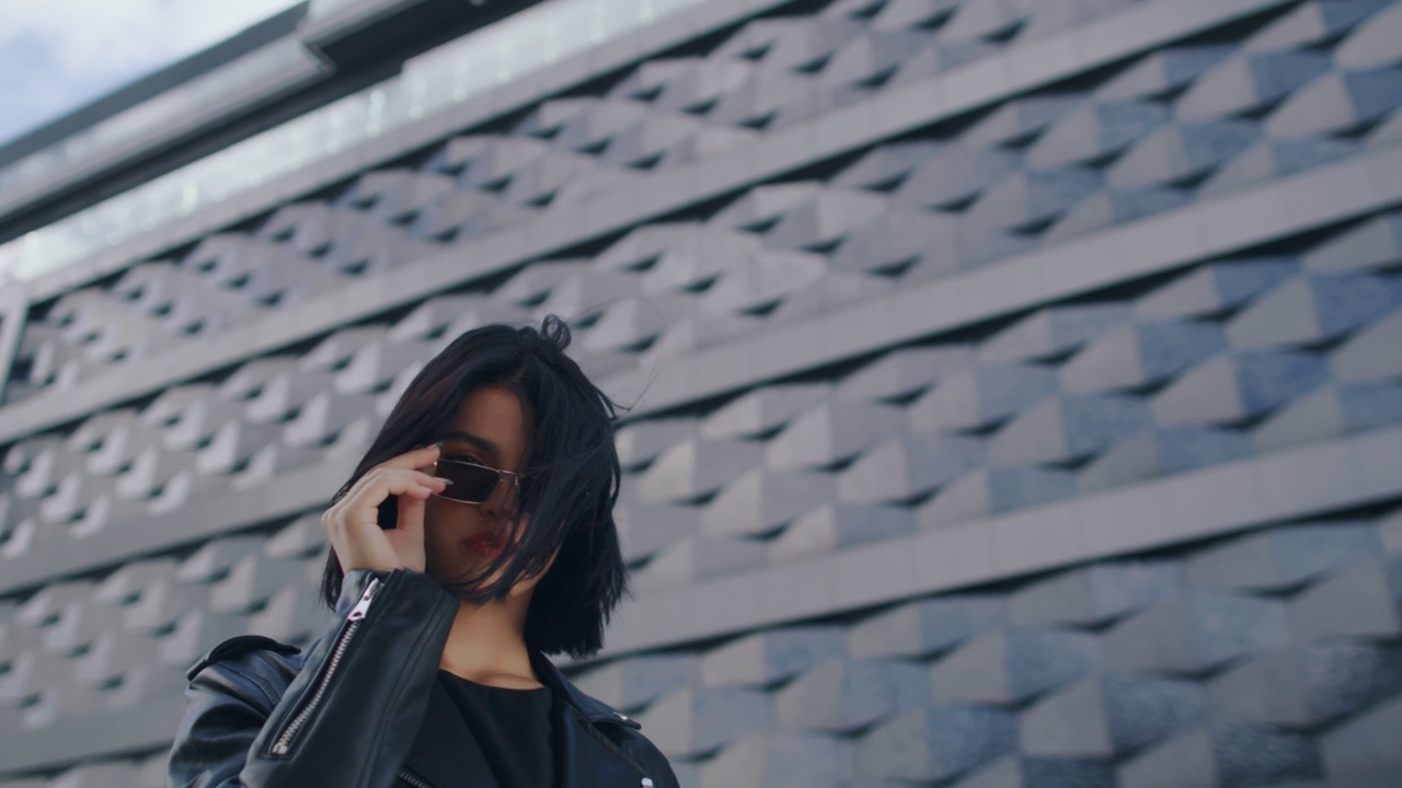 自信的年轻黑发女子在时尚的外观，穿着黑色皮夹克和太阳镜看着相机站在城市街道的大楼，触摸她的头发。生活方式,时尚,城市视频素材