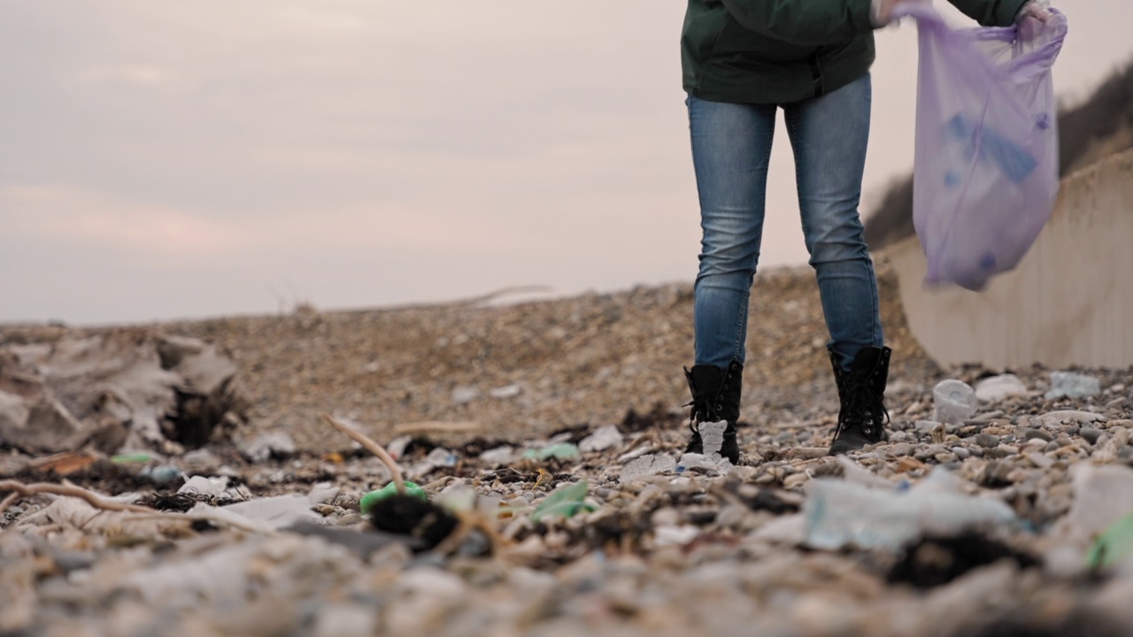 地球日。一名女性志愿者在海滩上收集垃圾，并将其放入垃圾袋。低角度的观点。环境污染的概念视频素材