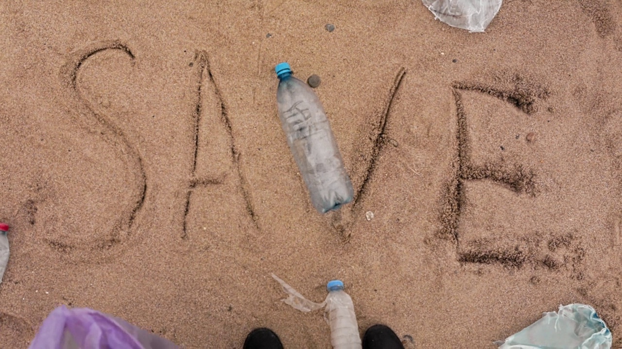 地球日。志愿者在沙滩上捡起塑料瓶，将垃圾放入垃圾袋。文字保存在沙子上。前视图。59、环保与污染的理念视频素材
