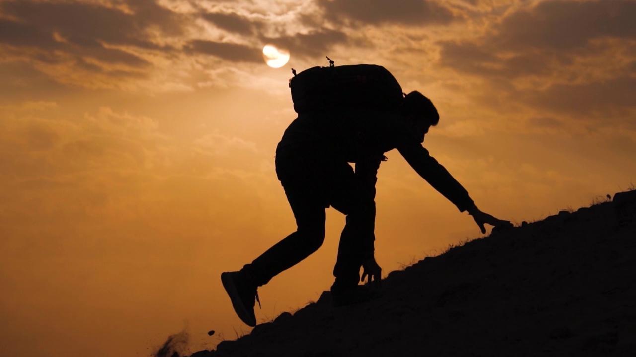 一个徒步旅行者的剪影拍摄在太阳前面的陡峭的山在日落与云。登山者挣扎着爬上顶峰，却滑倒了。冒险的背景。视频下载