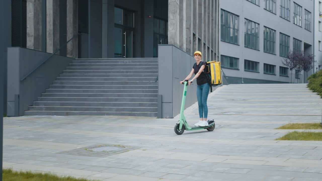 微笑着送食物的女快递员背着黄色保暖背包，骑着电动滑板车走在街上。积极的送货员，在线客户订单送货视频素材