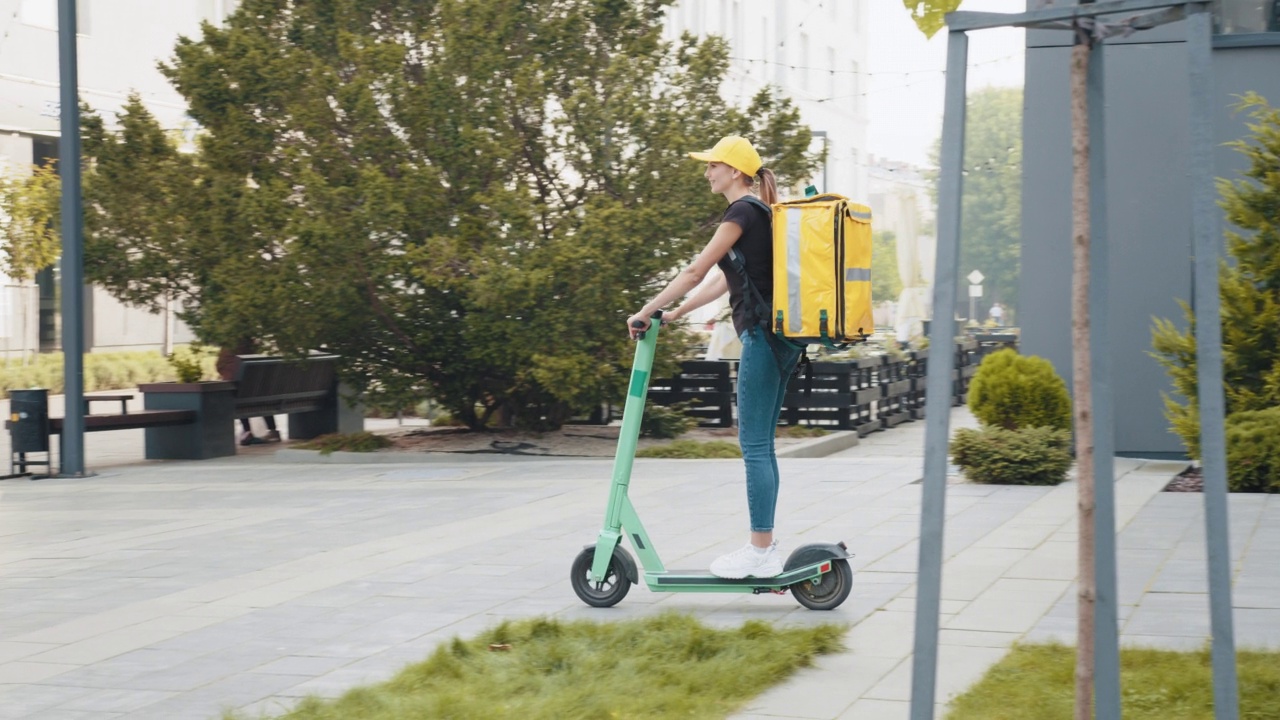 年轻的快递员骑着他的电动滑板车。快递员骑着一辆电动摩托车，背着黄色背包走在空荡荡的人行道上。商品的交付视频素材