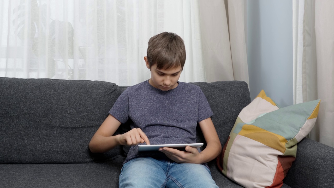 十几岁的男孩坐在家里的沙发上使用数码平板电脑。孩子使用应用程序学习，玩游戏，上网，看视频，在线学习视频素材