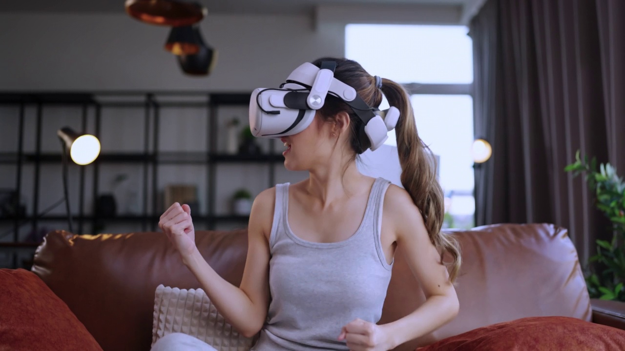 年轻迷人的休闲亚洲女性通过VR眼镜头戴护目镜在沙发上观看了虚拟现实中的表演直播音乐会，在家里的客厅里享受着乐趣和快乐的时刻视频下载