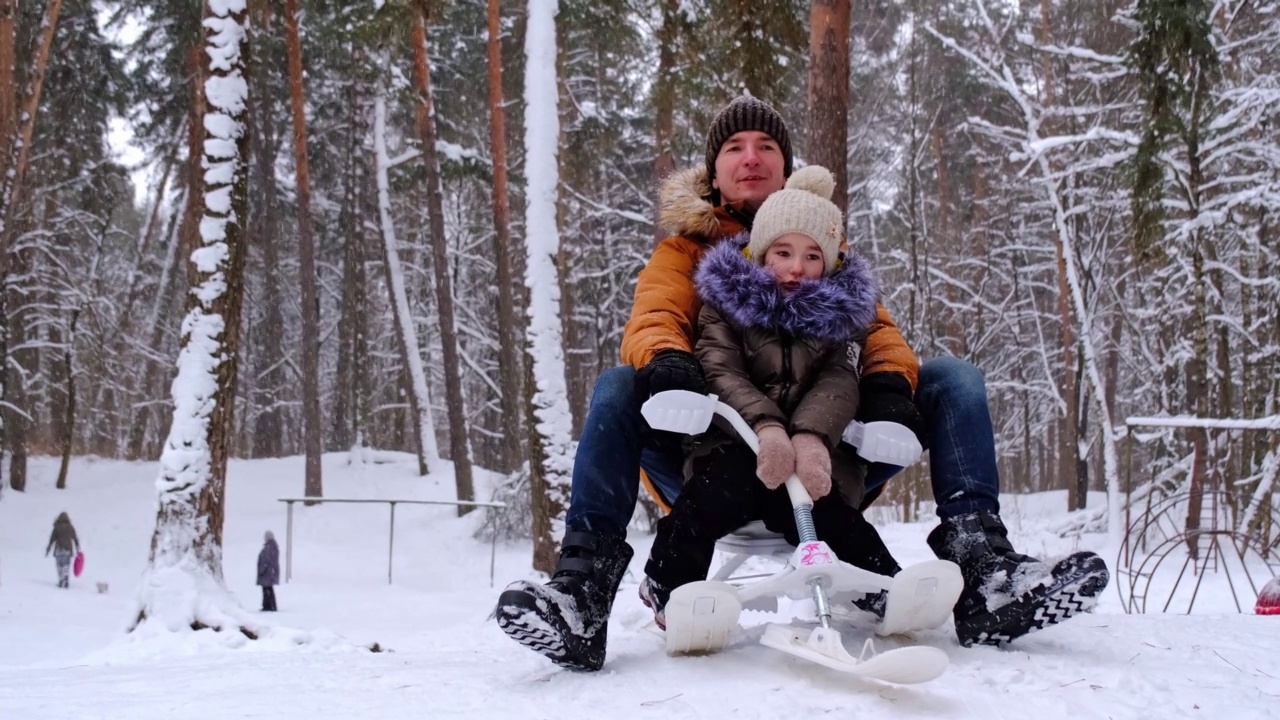 在一个下雪的冬天，爸爸和女儿骑着雪车下山。他们坐在同一个雪橇上。冬季家庭娱乐，情绪高涨视频素材