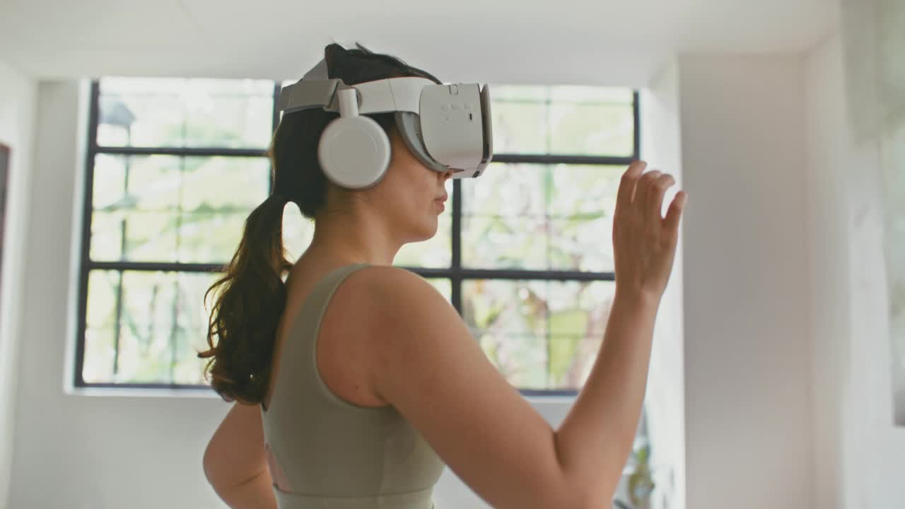 年轻的运动女性戴上VR头盔，在家里的客厅锻炼体验模拟数字世界视频素材