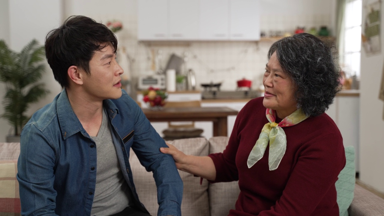 快乐的亚洲母亲拥抱她的成年儿子，他来拜访她在家里的客厅。久别重逢后，他们在沙发上愉快地聊天视频下载