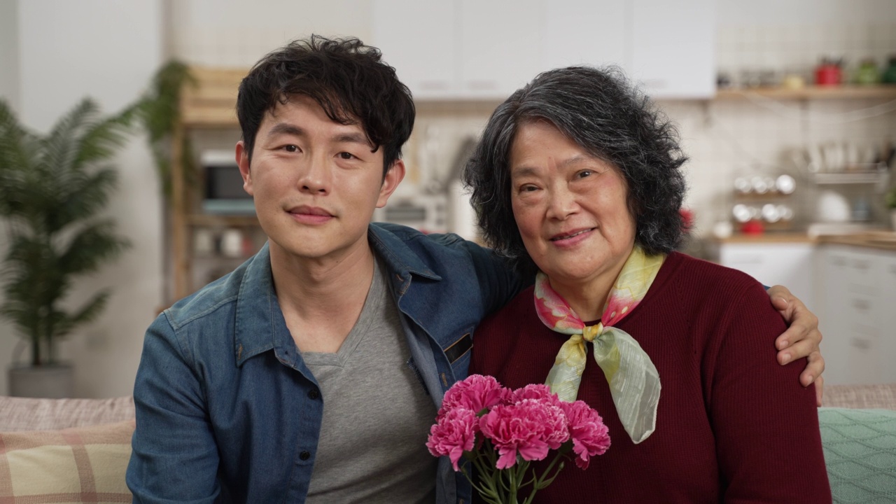 在家里的客厅里，亚洲母亲和成年儿子带着康乃馨看着相机微笑的照片。代爱和母亲节的理念视频下载