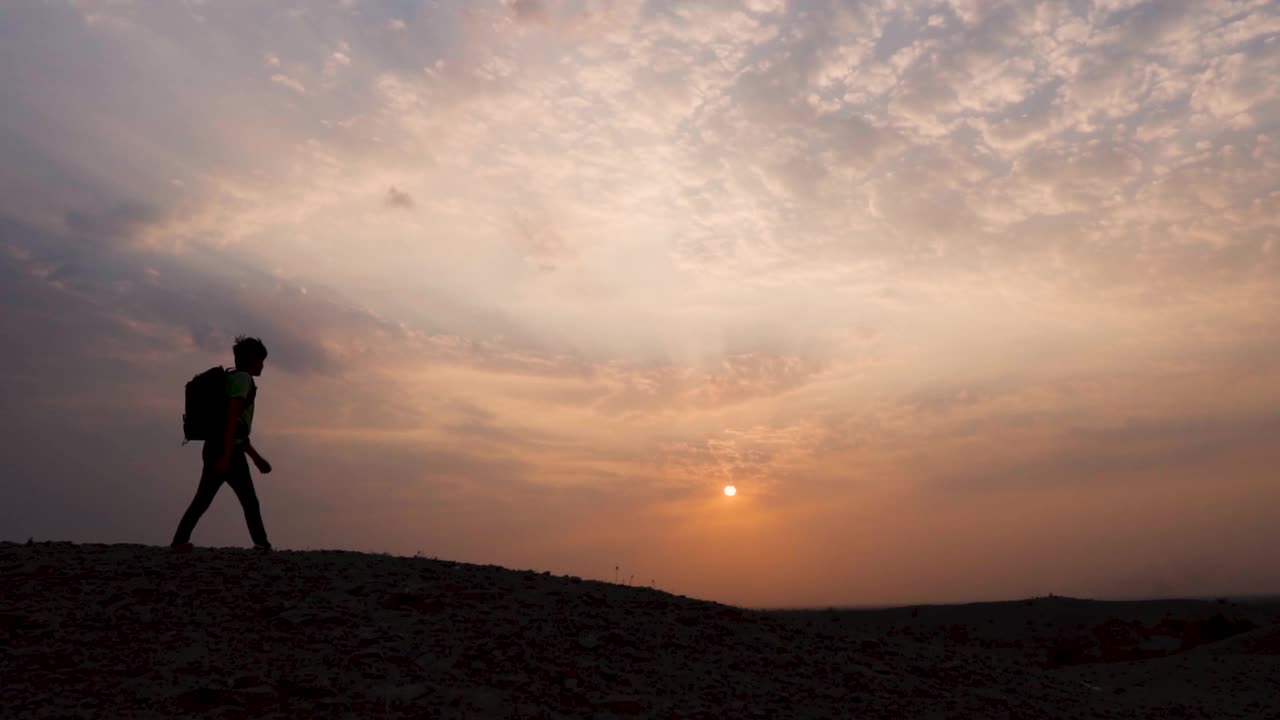 一个印度徒步旅行者在日落前的山顶上行走的剪影。自然背景。日落时分，一个人走在橙色的云彩和太阳前面。视频素材