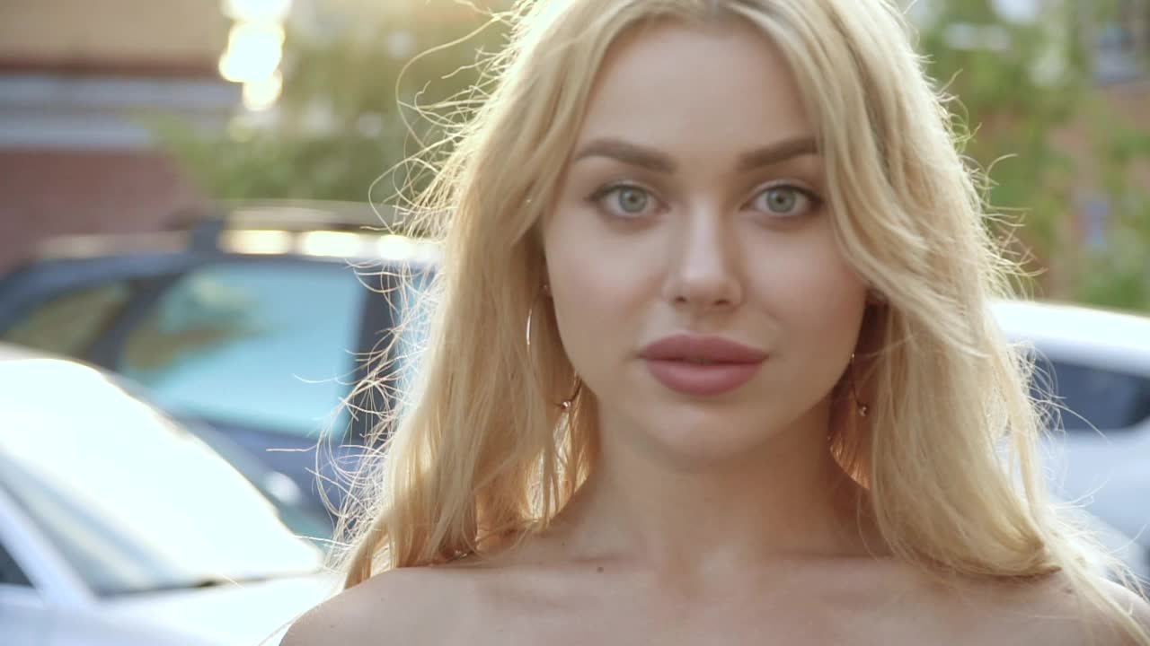 一个丰满的嘴唇和蓝眼睛的金发美女的肖像在停车场的背景上视频下载