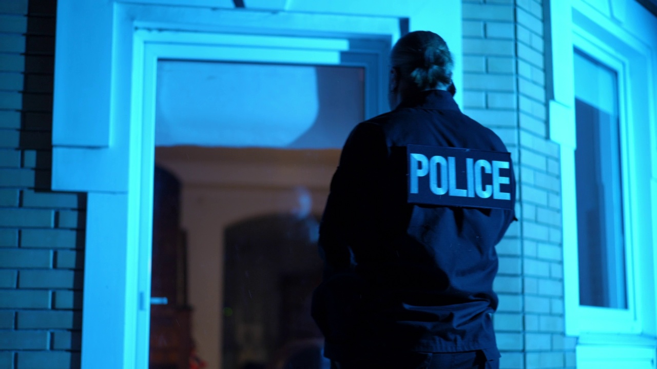 一名身穿制服的巡警站在黑暗的房子前打电话，警灯闪烁。白人警察在犯罪现场的中景镜头。视频下载