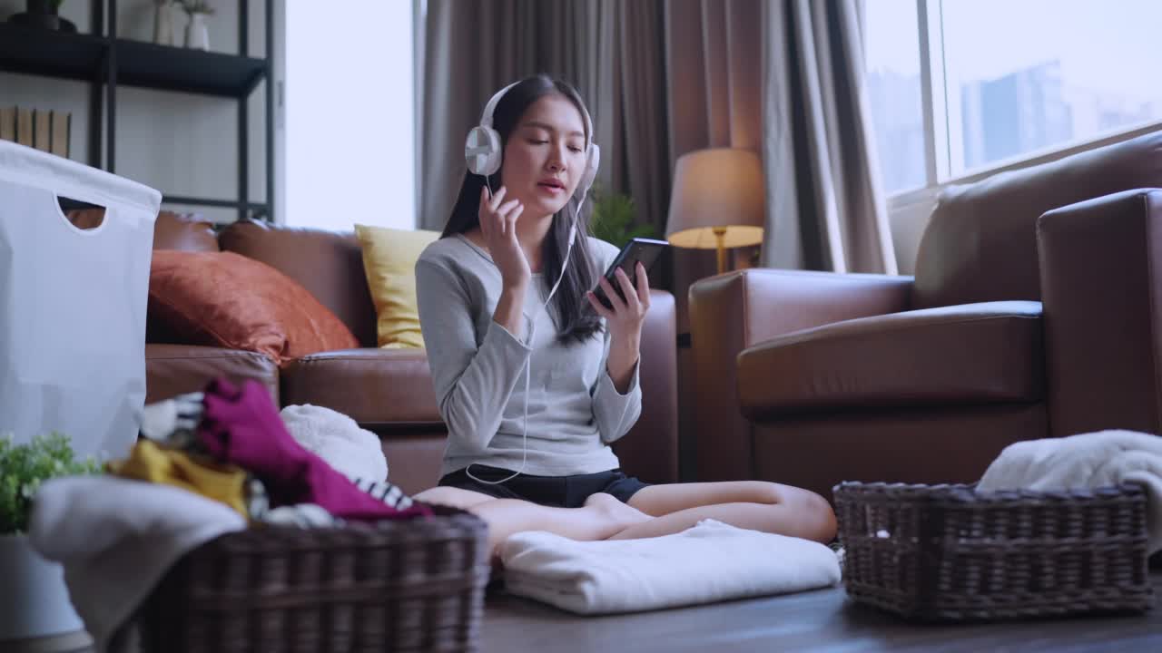 周末，亚洲女性家庭主妇一边叠衣服，一边从智能手机上挑选音乐播放列表视频下载