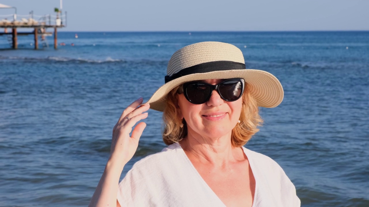 快乐的50岁的成熟女人穿着白色的连衣裙，在海滩附近的沙滩上的稻草沙滩和太阳镜。度假观念，放松，退休年龄视频素材
