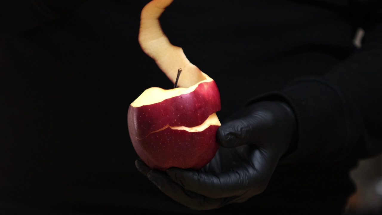 戴着黑色手套的人手清洁红苹果(螺旋状)。厨师手削苹果。在黑色背景上稍微削了皮的苹果。多汁的苹果正在剥皮视频下载