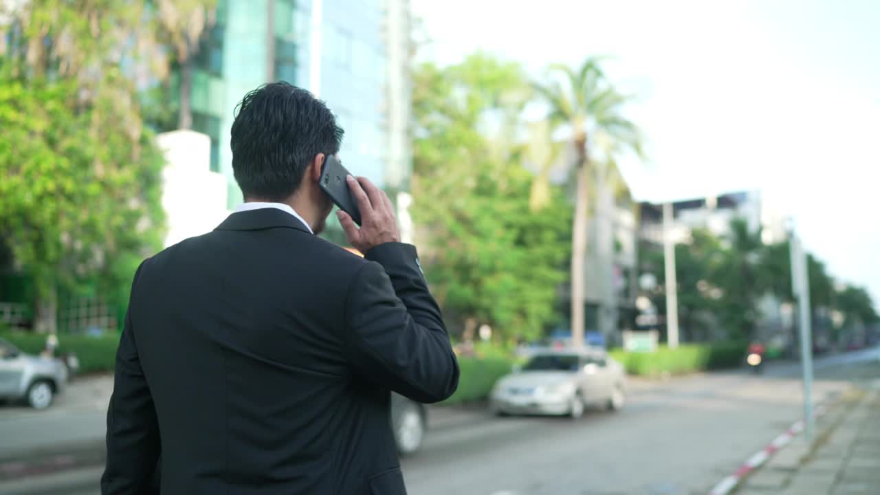一个成功的商人走在街上，手里拿着一部电话。一个在户外使用智能手机的自信男人。一个开心的商人正在外面用手机。视频素材