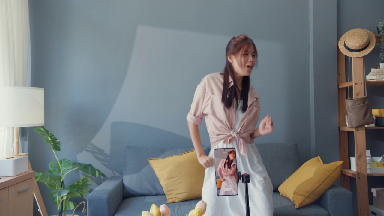 快乐的年轻亚洲女孩博主在家里的客厅里用手机摄像头录制视频和舞蹈内容。社会距离冠状病毒大流行概念。自由和积极的生活理念视频下载
