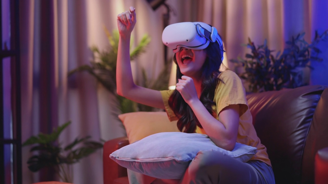 年轻迷人的休闲亚洲女性通过VR眼镜头戴护目镜在沙发上观看了虚拟现实中的表演直播音乐会，在家里的客厅里享受着乐趣和快乐的时刻视频下载