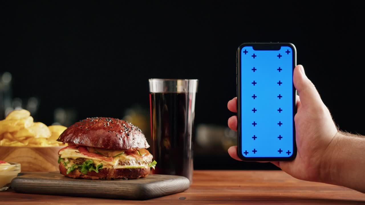 美味的汉堡和智能手机与蓝色色度键屏幕特写，煮多汁的三明治，快餐配送应用程序，准备自制的肉和新鲜蔬菜汉堡食谱。视频素材