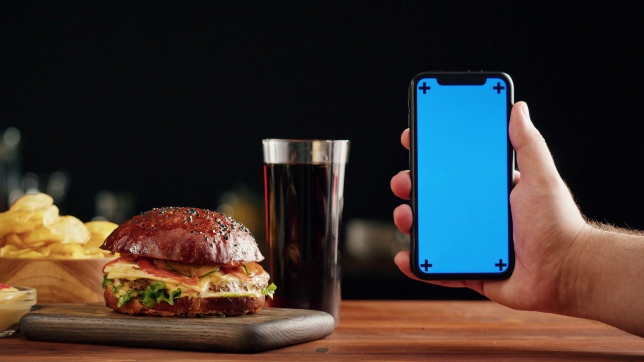 美味的汉堡和智能手机与蓝色色度键屏幕特写，煮多汁的三明治，快餐配送应用程序，准备自制的肉和新鲜蔬菜汉堡食谱。视频素材