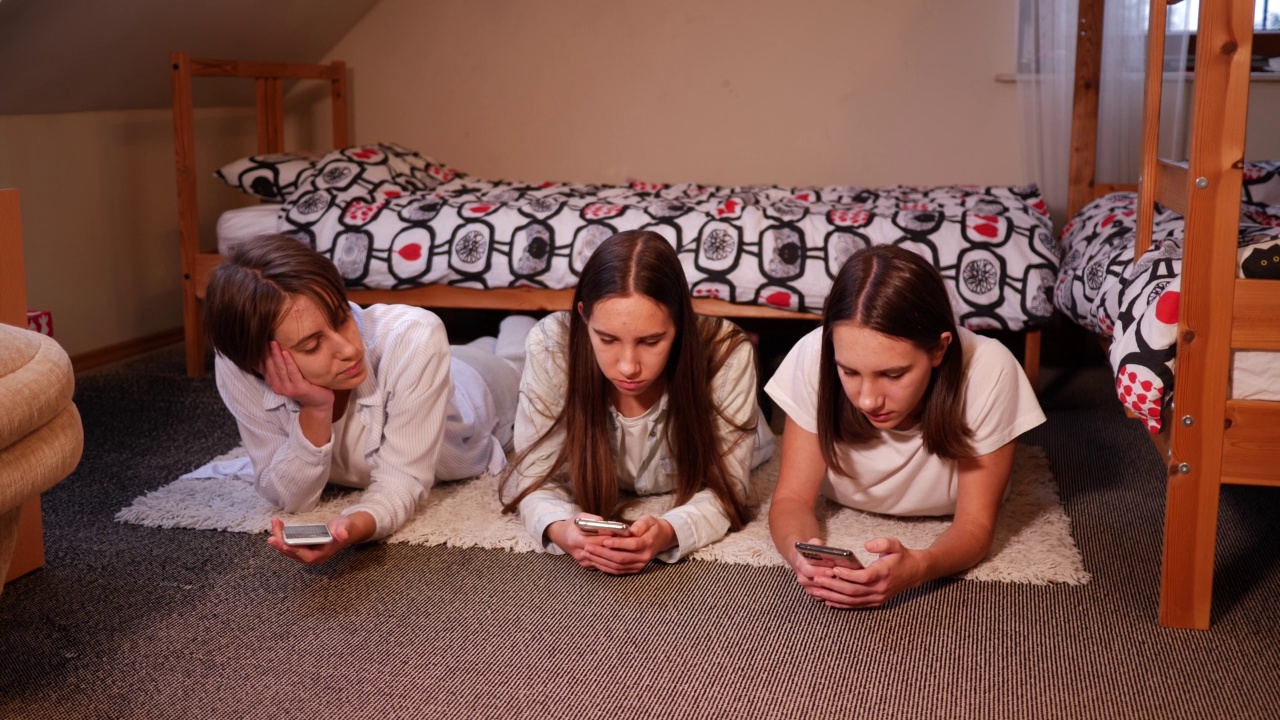 三姐妹十几岁的女孩在家里使用智能手机，躺在卧室的地板上，花时间在社交媒体视频素材