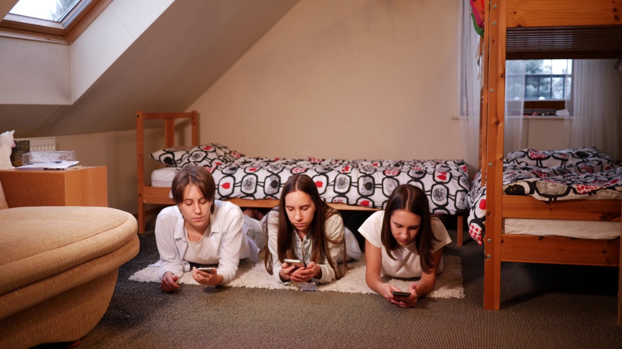 三姐妹十几岁的女孩在家里使用智能手机，躺在卧室的地板上，花时间在社交媒体视频素材