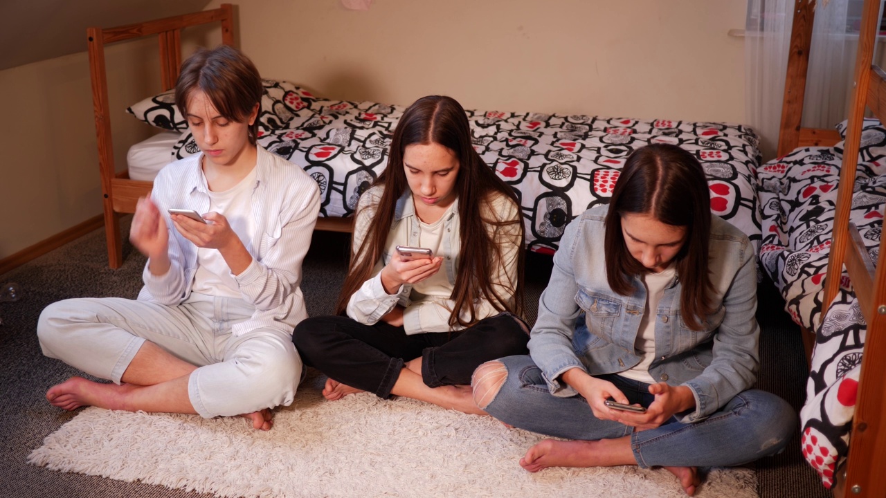 三姐妹十几岁的女孩在家使用智能手机，坐在卧室的地板上，花时间在社交媒体视频素材