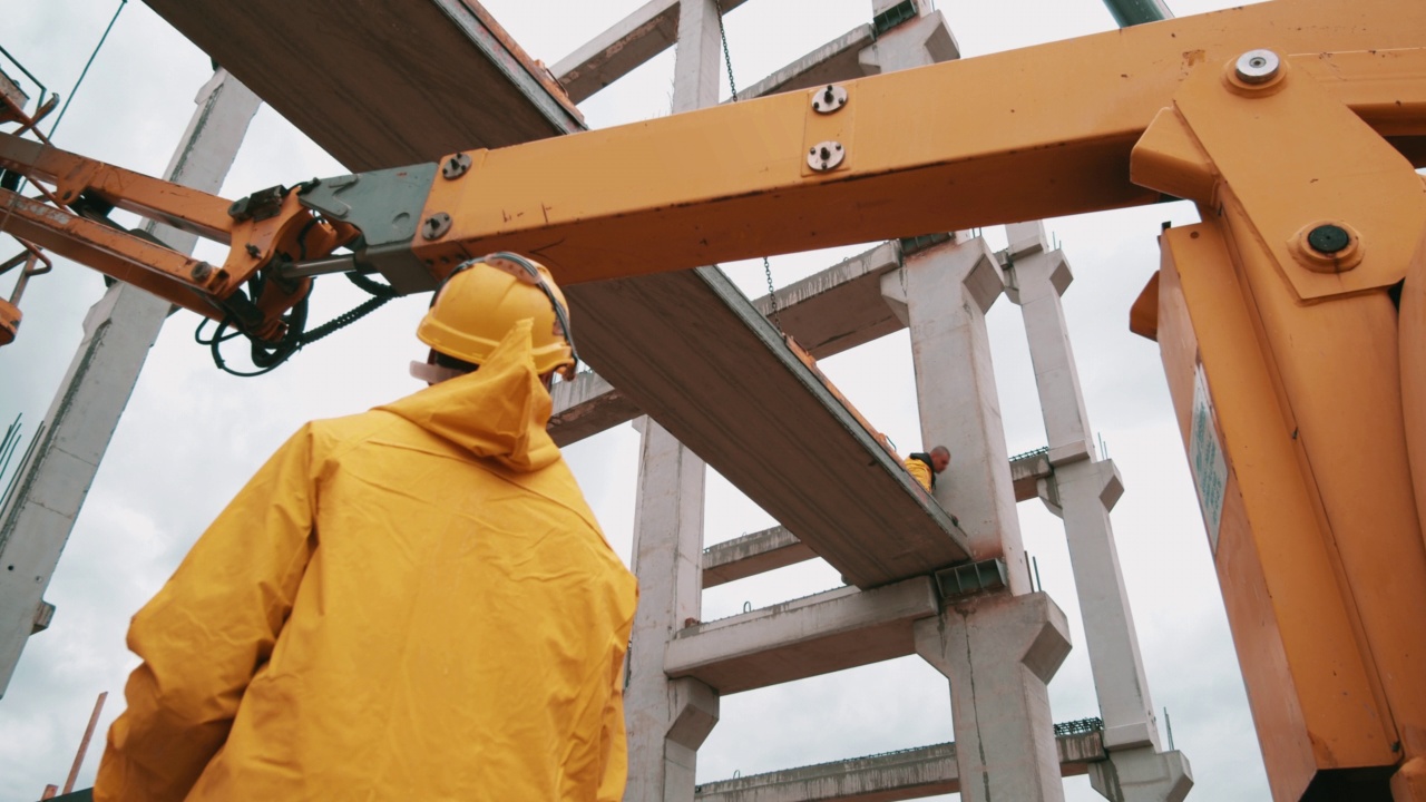 身穿黄色雨衣的建筑工人们正在工地上竖起巨大的混凝土柱子。用起重机和空中平台安装预制构件。股票视频。视频素材