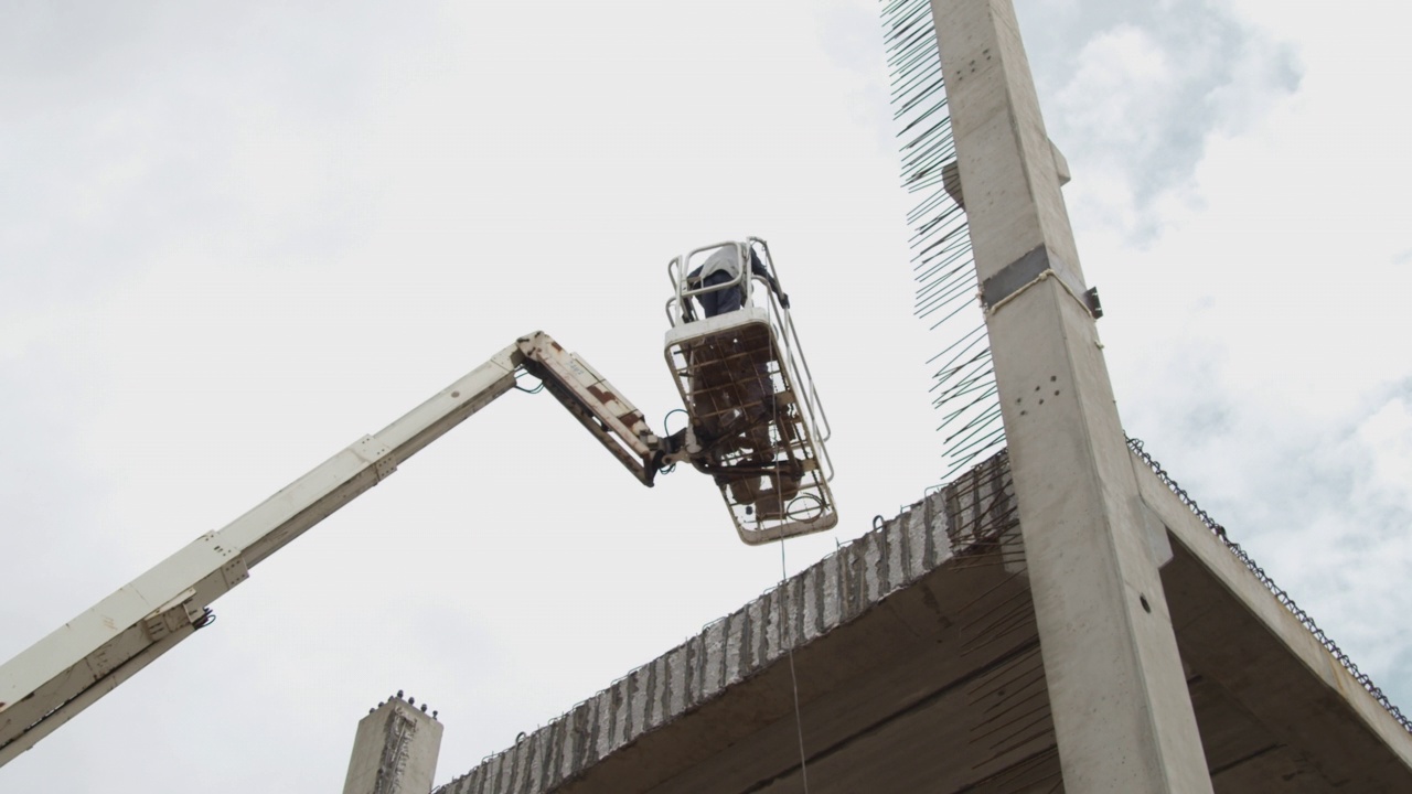 建筑工人在吊车篮里，悬挂在由大型预制构件组成的建筑结构上。建筑工人们正在竖混凝土柱子。在他们的身后是阴沉的天空。股票视频。视频素材