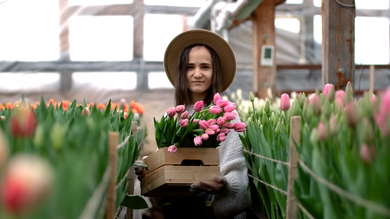 一个戴着帽子的年轻农家女孩拿着一个装着郁金香花束的木箱。温室里的白人女商人视频素材