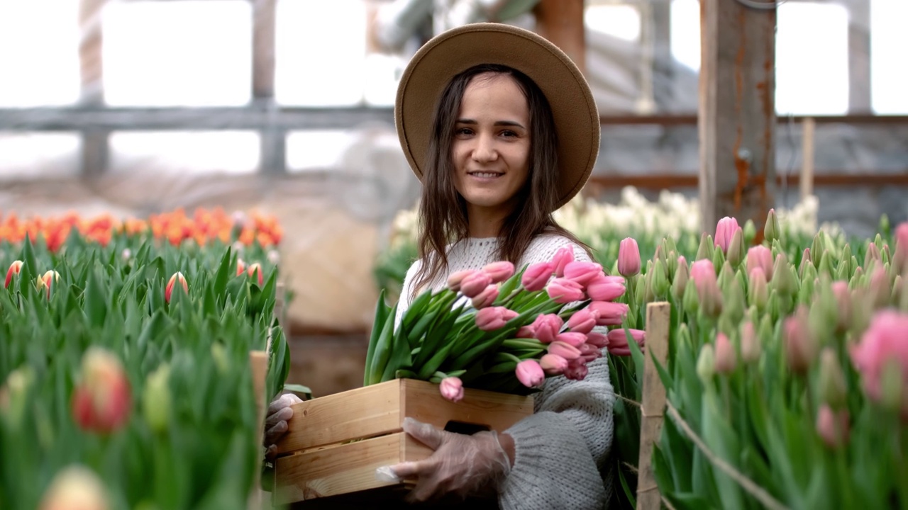 一个年轻的农民女孩戴着帽子，手里拿着一个盒子，里面放着郁金香花束。温室里的白人女商人视频素材