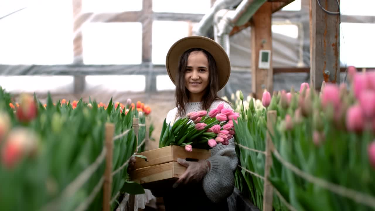 一个年轻的农民女孩戴着帽子，手里拿着一个盒子，里面放着郁金香花束。温室里的白人女商人视频素材