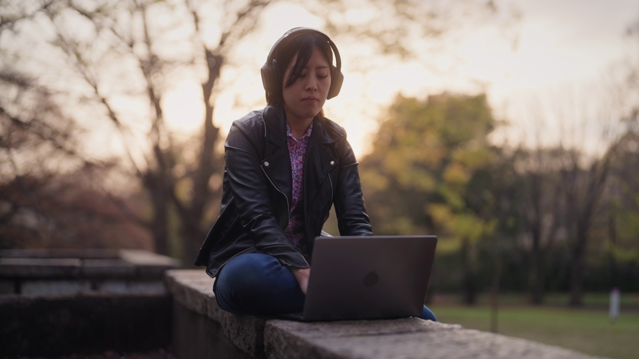 年轻女子坐在公园里，在日落时使用笔记本电脑视频下载