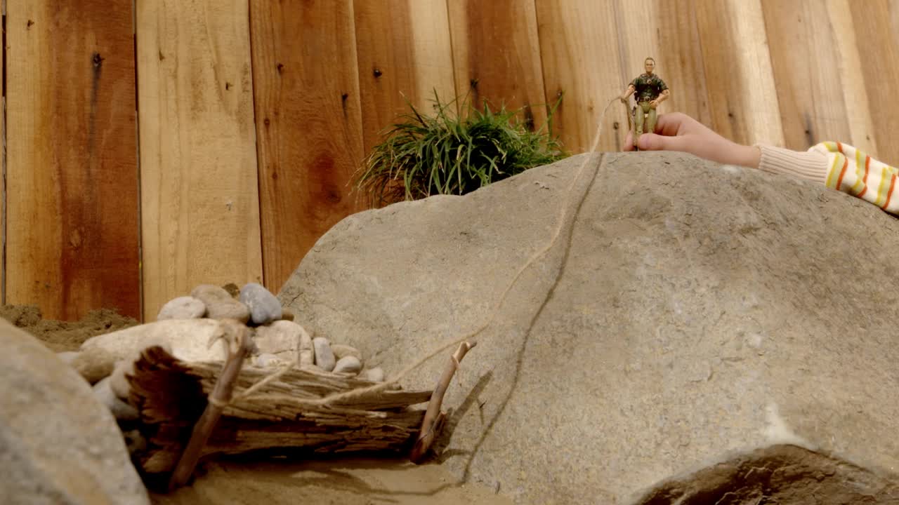 孩子的手在岩石上玩动作玩偶玩具的特写角度。可能是后院。动作玩偶将绳子系在自制的大坝上，可以看到岩石。在bg可见的木栅栏。视频下载