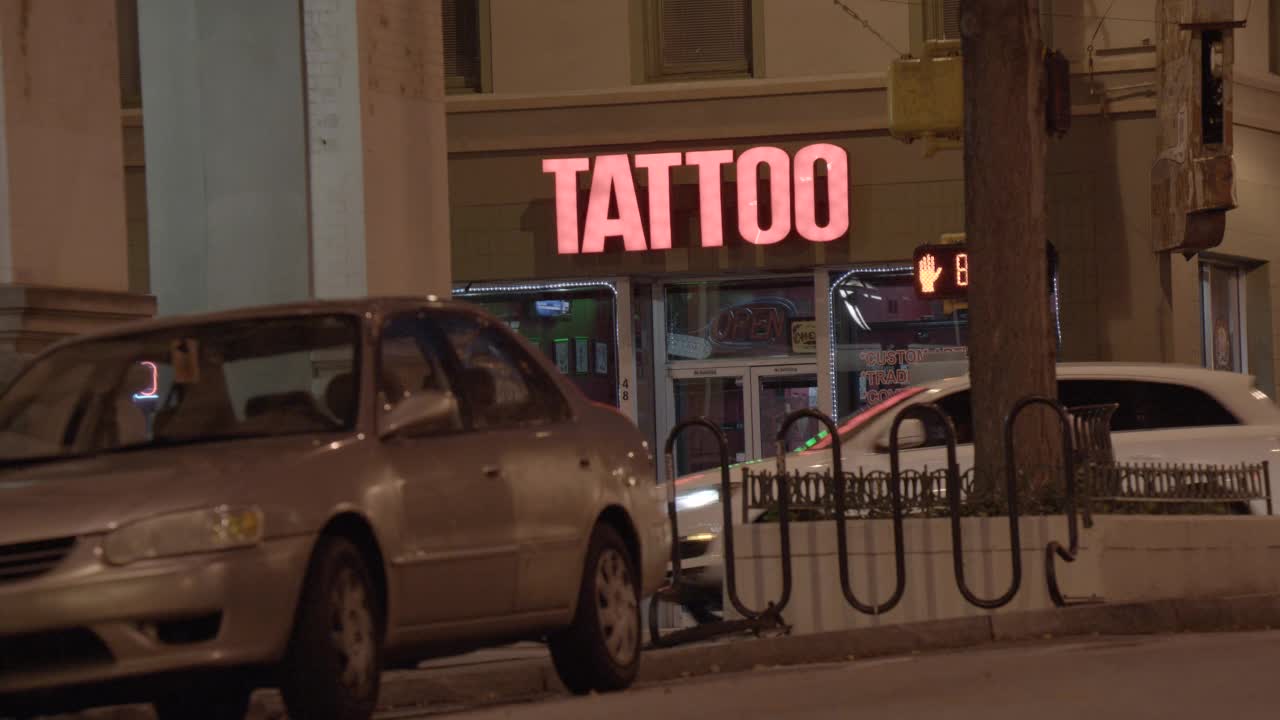刺青店或商店入口霓虹灯的中角。可见车辆在城市街道上行驶和停放。行人清晰可见。视频素材