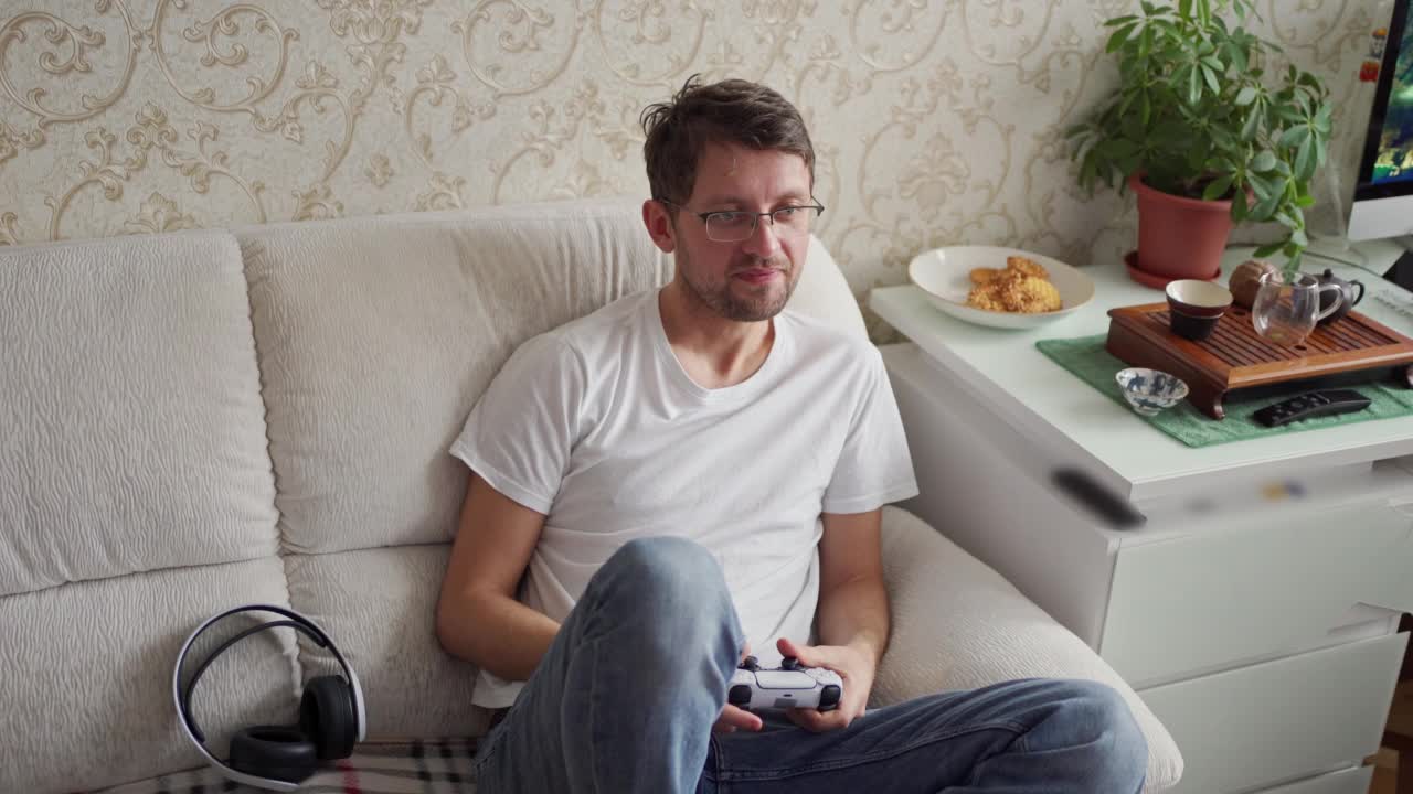 一个年轻人坐在沙发上玩游戏机。手握操纵杆视频下载
