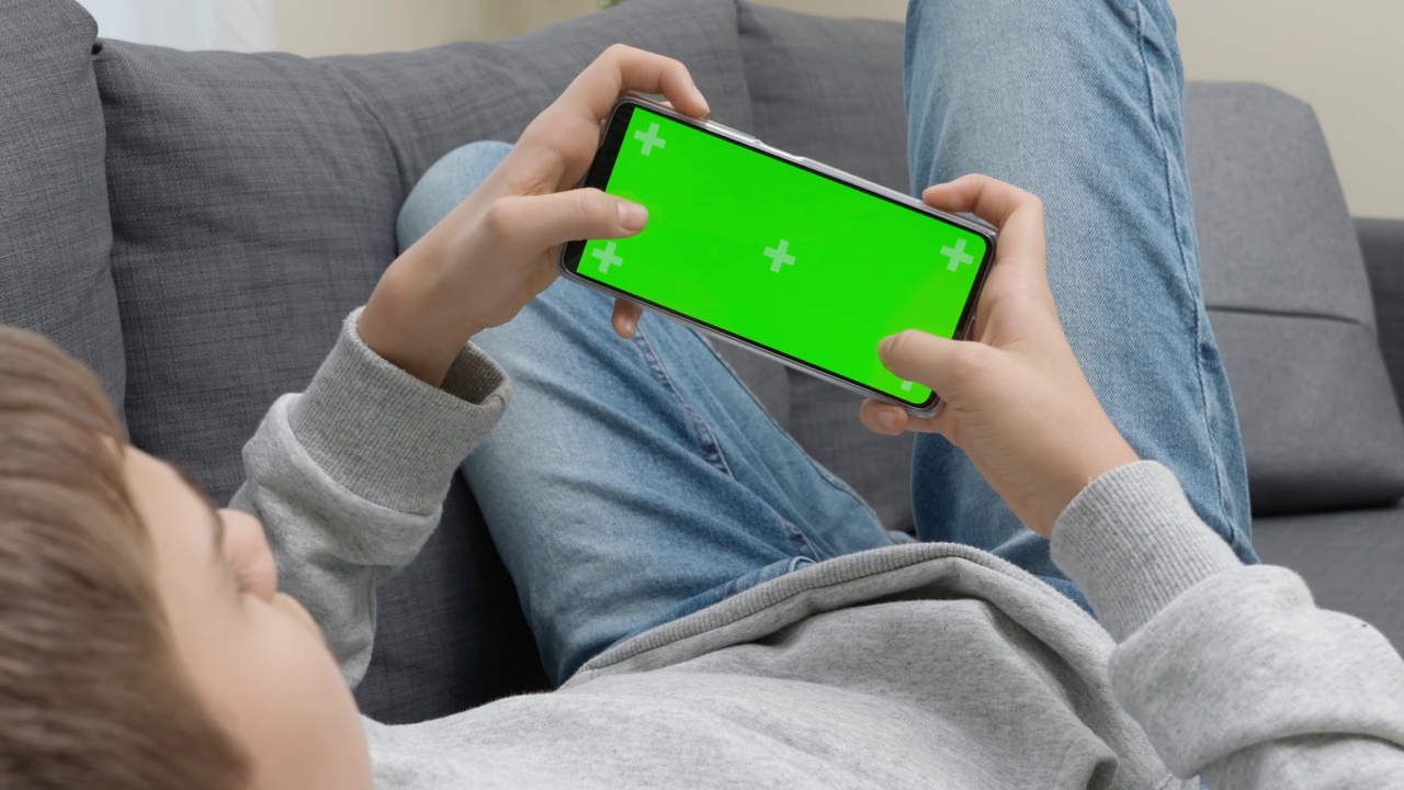 一个十几岁的男孩拿着手机在家里玩电子游戏。绿色背景上的手机屏幕。游戏成瘾，休闲，娱乐青少年视频素材