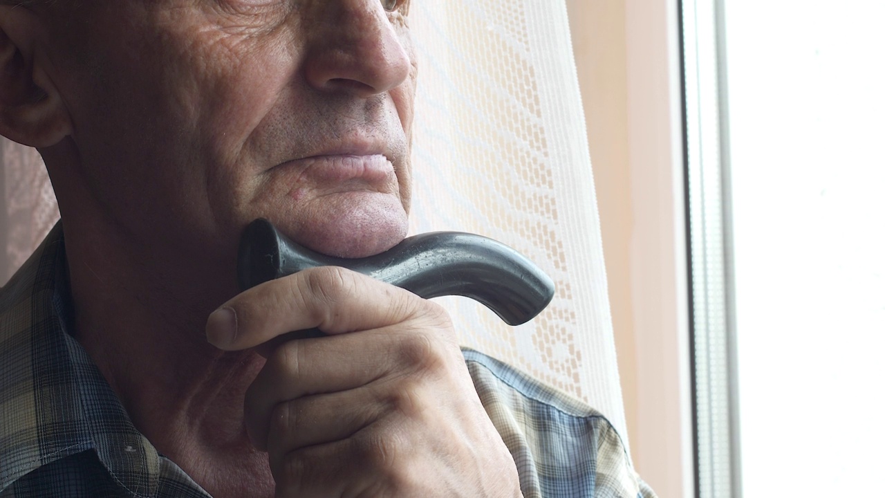 70多岁的白人老人拄着拐杖，表情严肃地望着窗外，等待着亲人。孤独的晚年主题。养老院。老人孤独的人视频素材