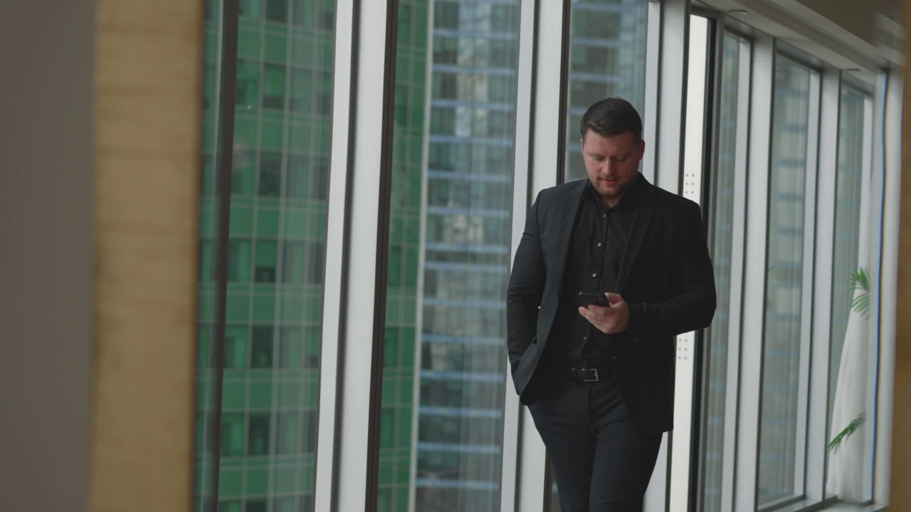一个商人在摩天大楼的背景下沿着全景窗户走着。英俊的商人在现代化的办公室里用手机打电话。商人正在用手机打电话视频下载