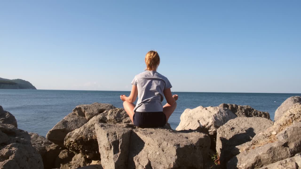 一个年轻女子坐在海边的莲花座上，看着黎明时分的大海。运动，冥想，平静，瑜伽，和谐，夏天。自然、健康、自由、个人活动的概念视频素材
