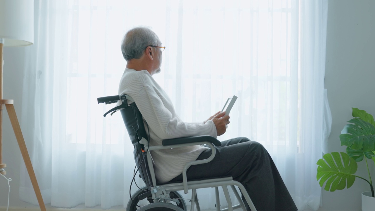 亚洲老年男性独自坐在轮椅上，看着家庭照片。年迈成熟的爷爷感到孤独和悲伤，在退休后呆在养老院想念他的亲戚和妻子。视频素材