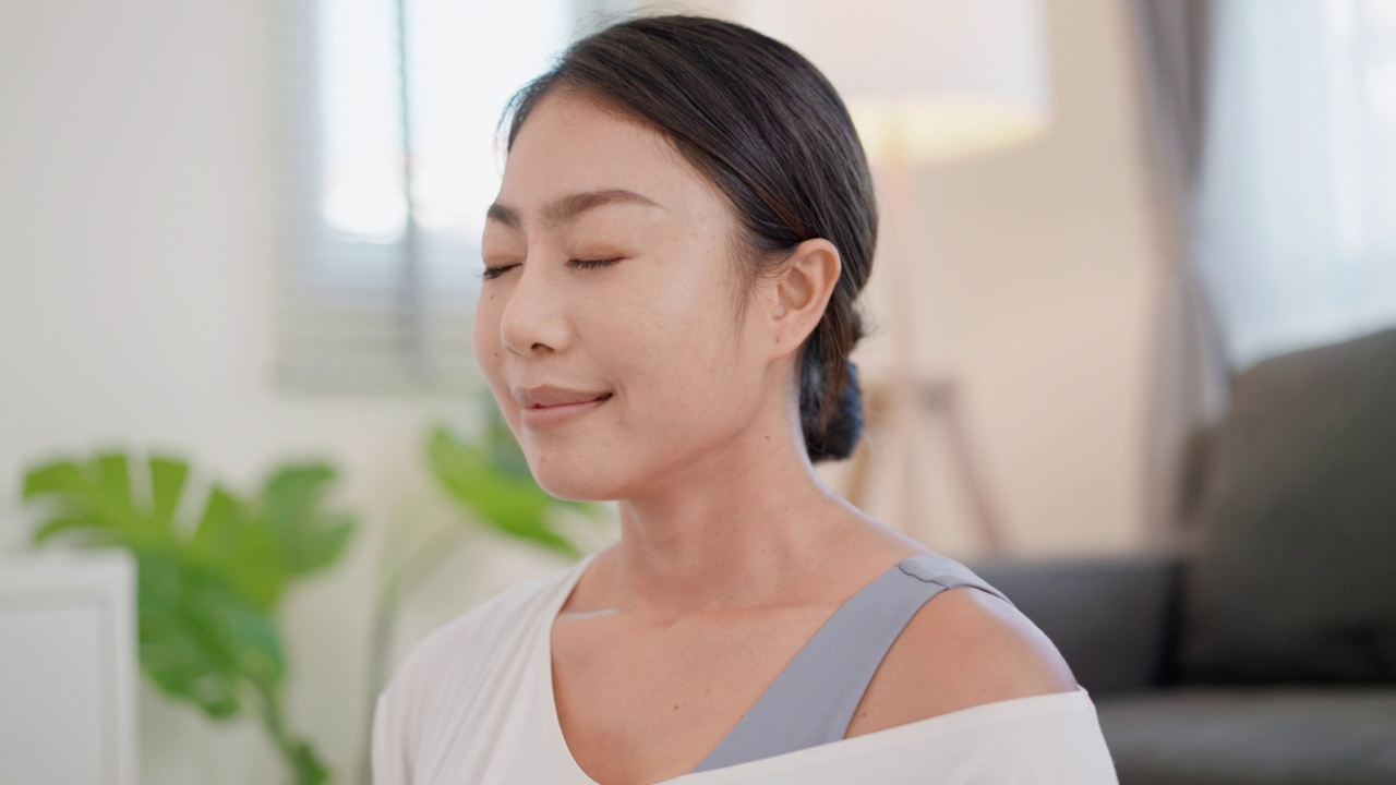 平静的亚洲女性穿着运动服坐在瑜伽床垫上，闭着眼睛深呼吸，冥想，专注于呼吸，在家里的客厅里以莲花姿势吸气和呼气。锻炼、休息、爱好、康乐活动的概念视频素材