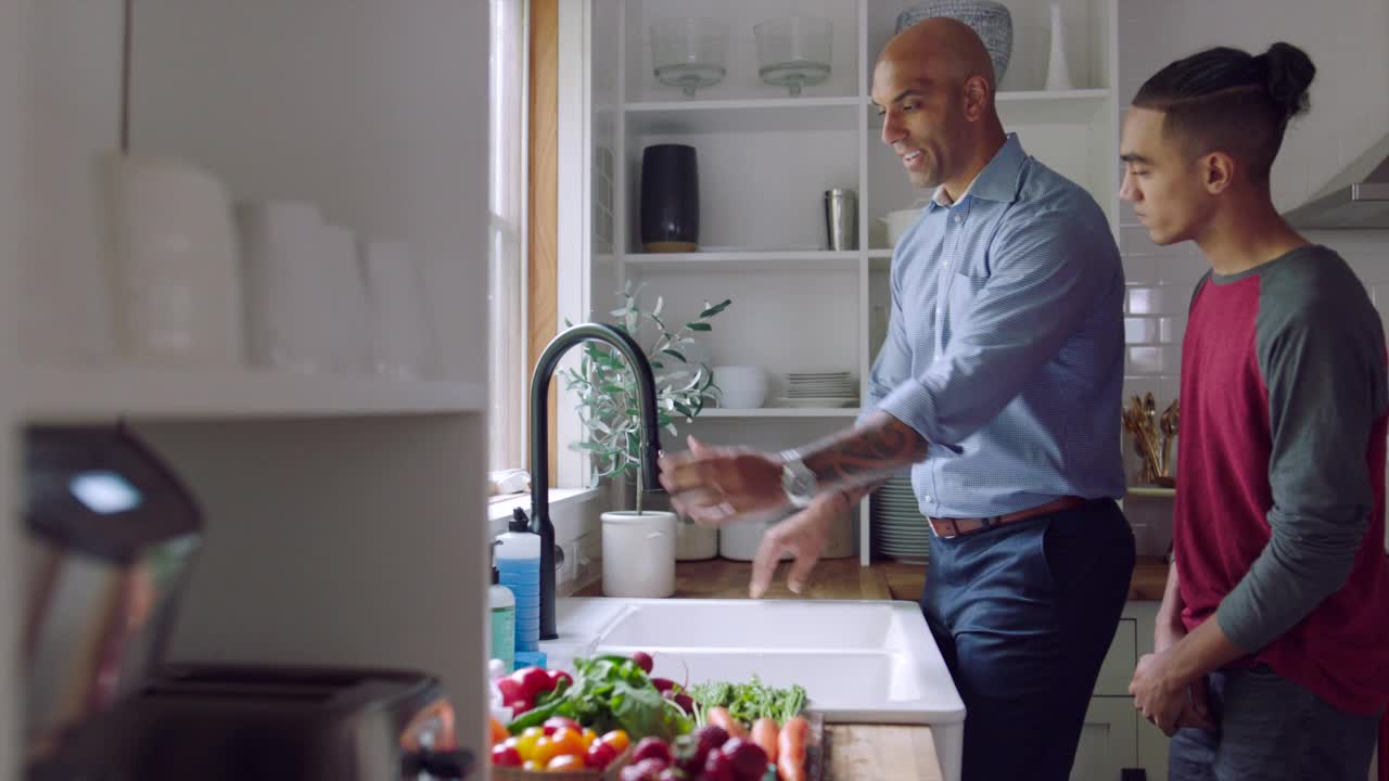 中宽镜头微笑的父亲和儿子洗新鲜蔬菜在厨房水槽视频素材