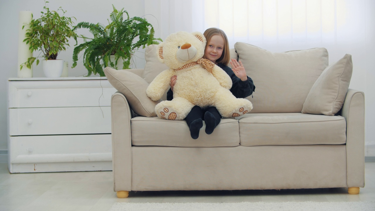 小微笑的女孩坐在沙发上她的玩具泰迪熊挥舞你好在4k慢动作视频。视频素材