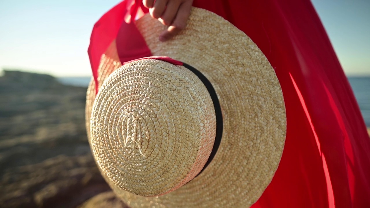 一名身穿红色连衣裙的年轻白人女子手持草帽，站在岩石嶙峋的海岸上。浅景深视频下载