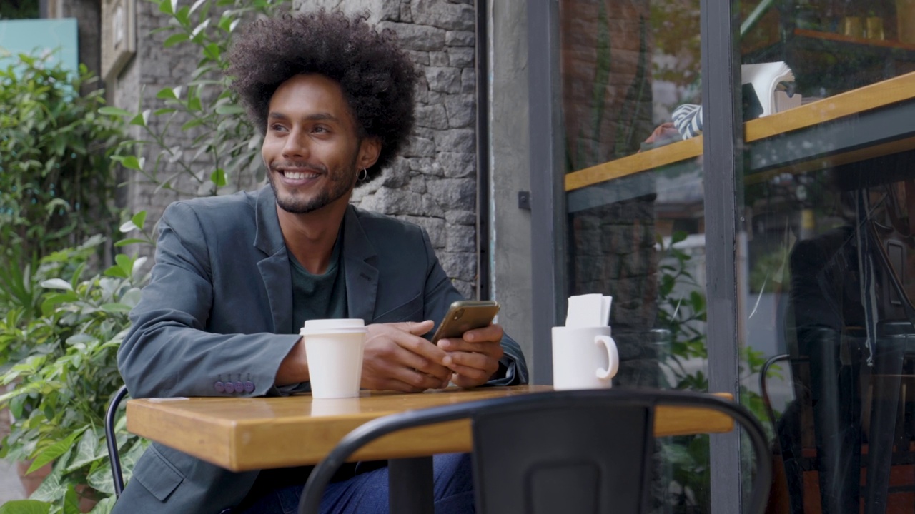 快乐的巴西人使用他的手机，而喝一杯咖啡在咖啡馆微笑视频素材
