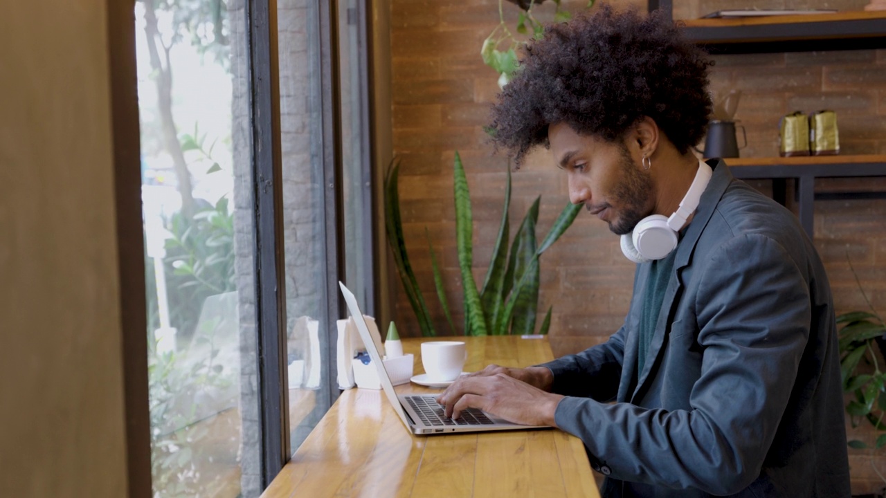 英俊的非洲裔美国人在咖啡店享受咖啡，然后结束休息，用笔记本电脑工作，戴着耳机专注于音乐视频素材