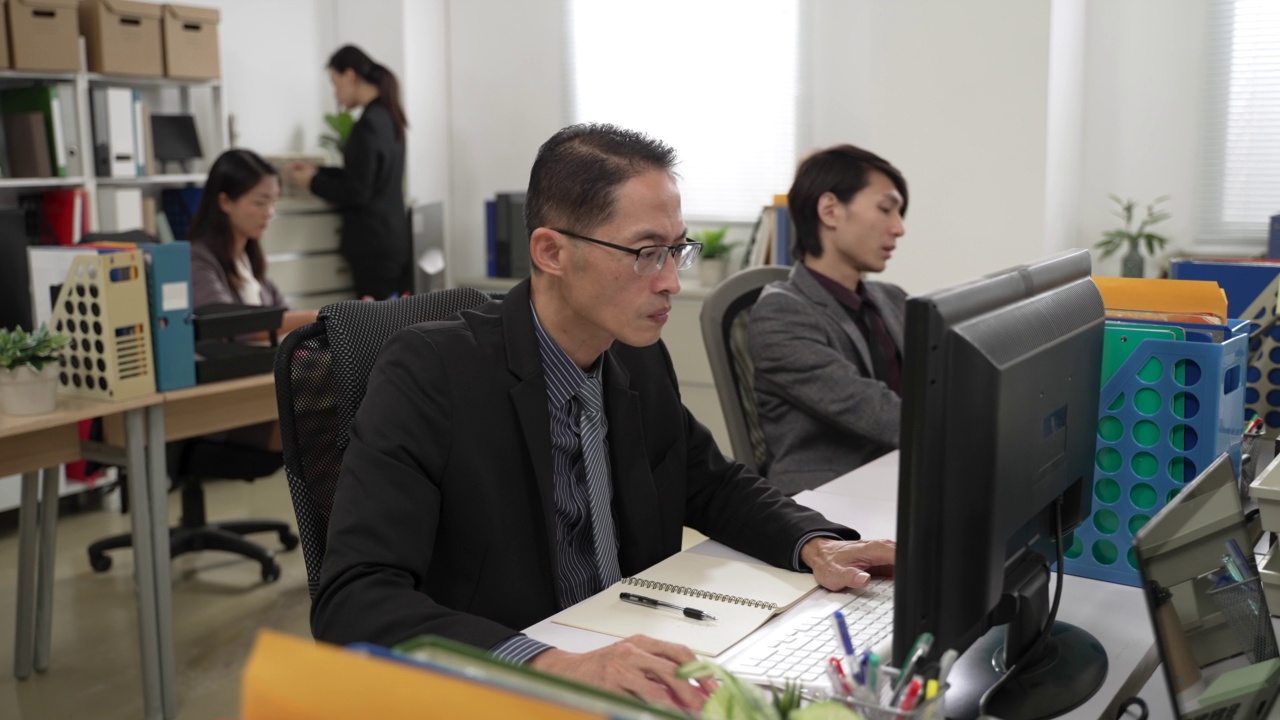 西装革履的亚洲商人在办公室与同事们聚精会神地在台式电脑上写笔记和工作视频下载